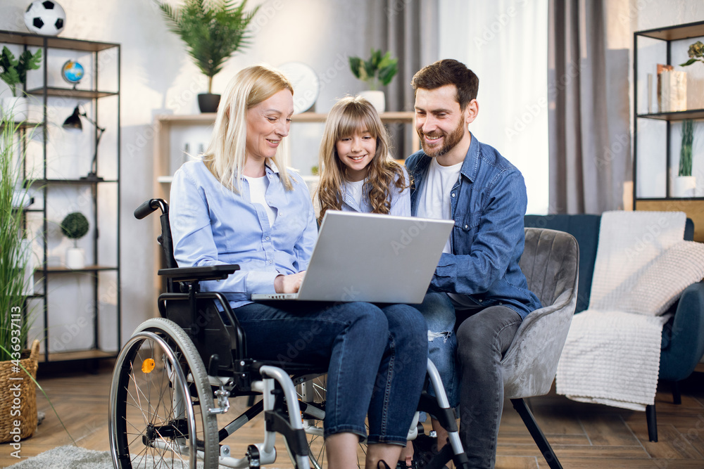 Handicapped woman with laptop on knees embracing her beloved daughter and husband. Happy young family enjoying time together at home. People with disabilities. Handicapped woman with laptop on knees embracing her beloved daughter and husband. Happy young family enjoying time together at home. People with disabilities.