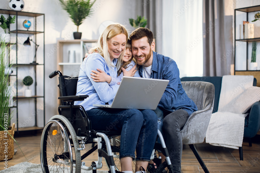 Handsome man and cute child hugging together woman in wheelchair with laptop on knees. Husband supporting beloved disabled wife. Family concept. Handsome man and cute child hugging together woman in wheelchair with laptop on knees. Husband supporting beloved disabled wife. Family concept.