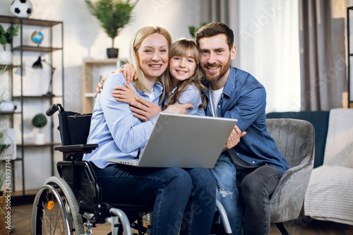 Handicapped woman with laptop on knees embracing her beloved daughter and husband. Happy young family enjoying time together at home. People with disabilities.