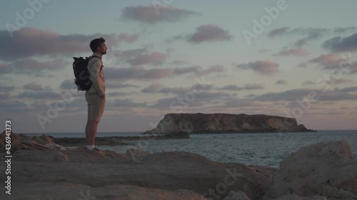 Man with backpack is hiking along a rocky coast at sunset near the Mediterranean sea in Cyprus. Male tourist admires the view Yeronisos, or Holy Island, lies off the coast of Agios Georgios Pegeias photo