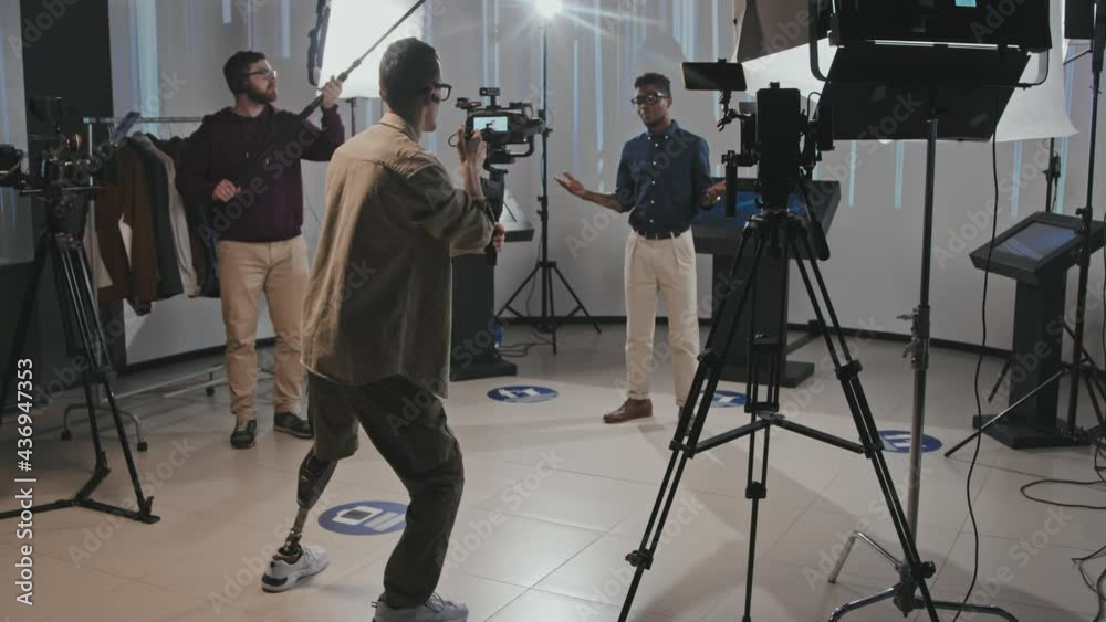 Full-shot slowmo of group of professionals with cameraman in head shooting video in studio. Man with leg prosthesis working as video operator walking with steady cam shooting video