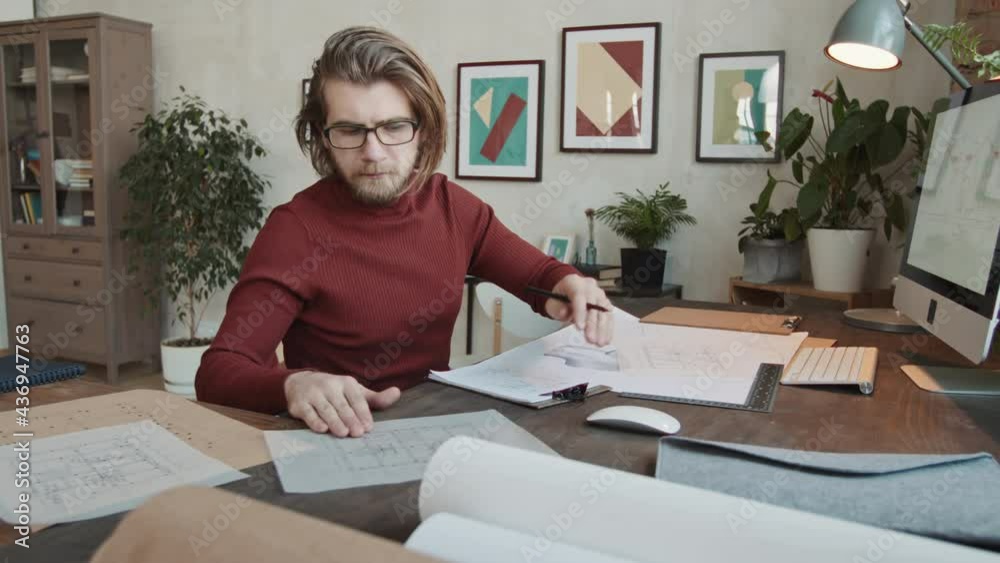 Medium slowmo of Caucasian male architect sitting in front of computer and drawing sketches of new house thinking of its design and layout Medium slowmo of Caucasian male architect sitting in front of computer and drawing sketches of new house thinking of its design and layout