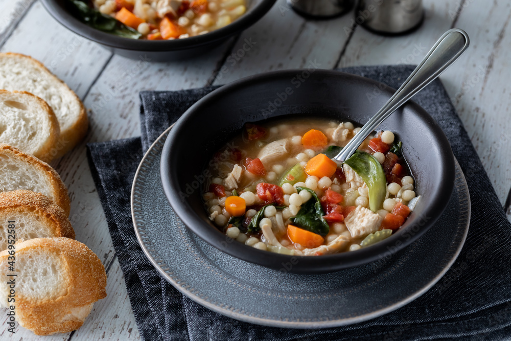 A close up look into a bowl of chicken vegetable and couscous soup, served with slices of fresh bread.