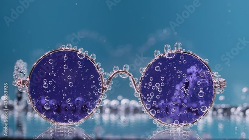 Round stylish sunglasses covered with air bubbles in transparent water on a blue blurred background. Slow motion. Close up. photo