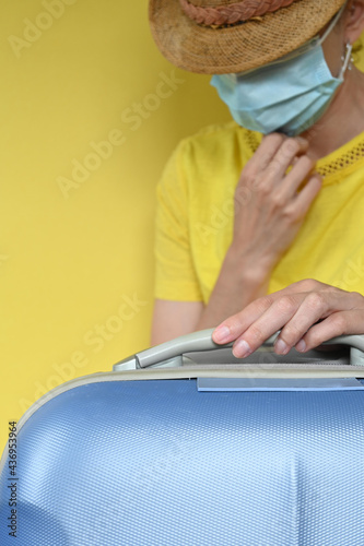 Vertical closeup of young latin woman wearing surgical mask  straw hat and yellow blouse and holding blue suitcase  yellow background with copy space