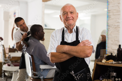 Portrait of professional aged barber standing with crossed arms in modern hair salon..