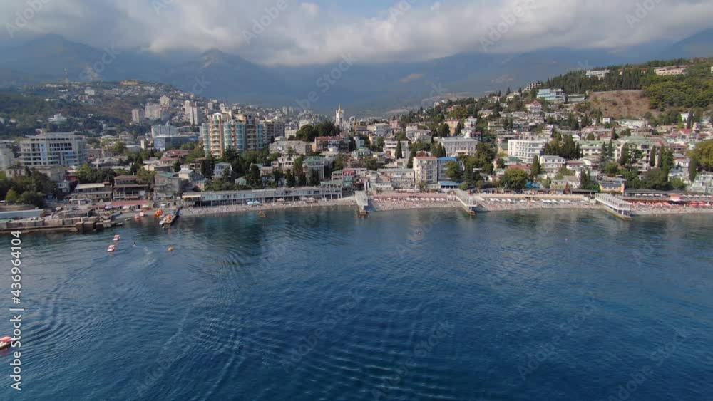 Bird’s eye view cinematic sunny summer city center seaside landscape Yalta Crimea Russia resort town. Black Sea, city beach, many people on shore. Modern hotels. Beautiful clouds. Side fast motion