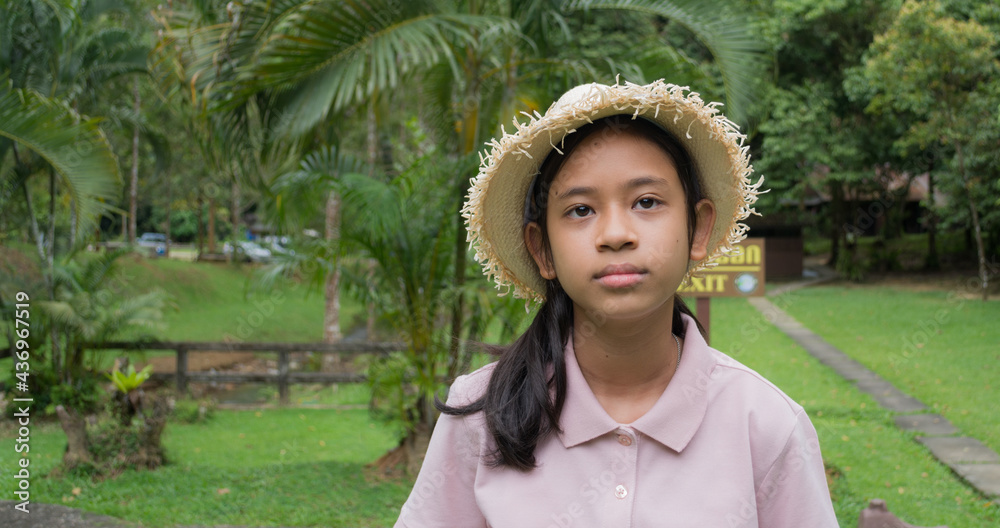 Portrait happy asian young girl wearing straw hat staying in forest park. Female youth volunteer guide standing on the lawn to welcome tourists in tropical forest.