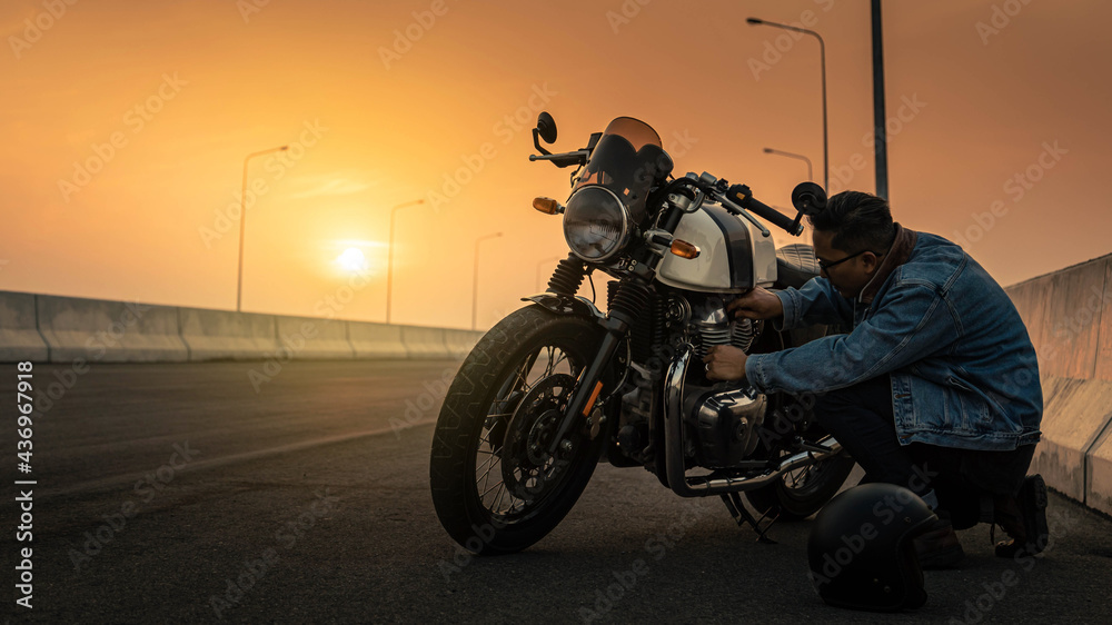 A man wearing a denim jacket is repairing a vintage motorcycle spark plug on a highway at sunset. Man riding a vintage motorcycle Biker Lifestyle Concept.