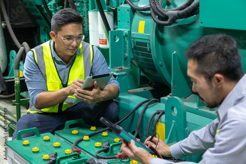 Two engineer working on checking and maintenance with batteries for diesel generator unit has a unit mounted radiator and fuel filter system at emergency power supply room.