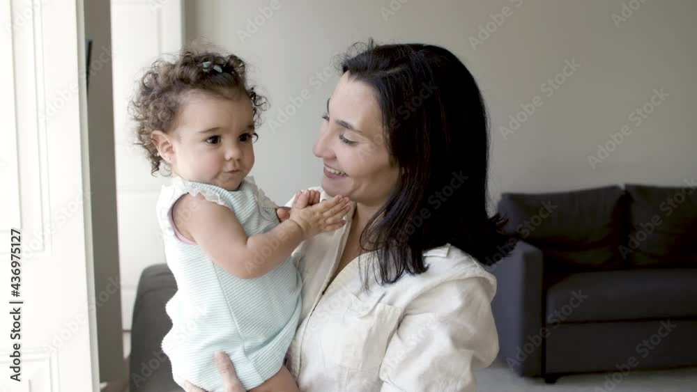 Single mother standing in living room, holding toddler in hands. Smiling Caucasian woman talking with her baby. Cute curly-haired girl hugging mother. Parenthood, infant, communication concept. Single mother standing in living room, holding toddler in hands. Smiling Caucasian woman talking with her baby. Cute curly-haired girl hugging mother. Parenthood, infant, communication concept.
