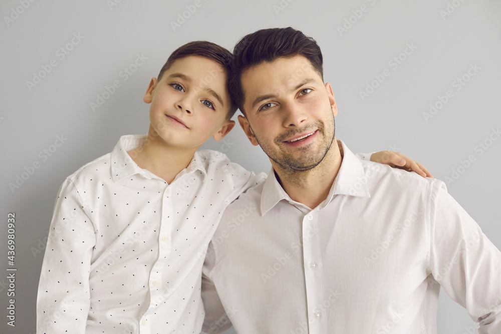 Close up portrait of father with son who are dressed in white shirts and cuddling while standing on gray background. Concept of family, good parents, love, happiness, childhood and fatherhood.