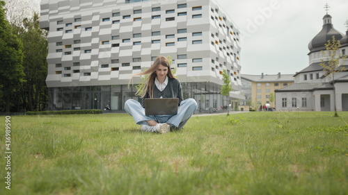 Busy attractive girl working at the laptop as sitting on grass in city park on hectic summer morning, outdoor shot in urban area. Student studying on computer sitting on campus staircase outside. © GRAFStock
