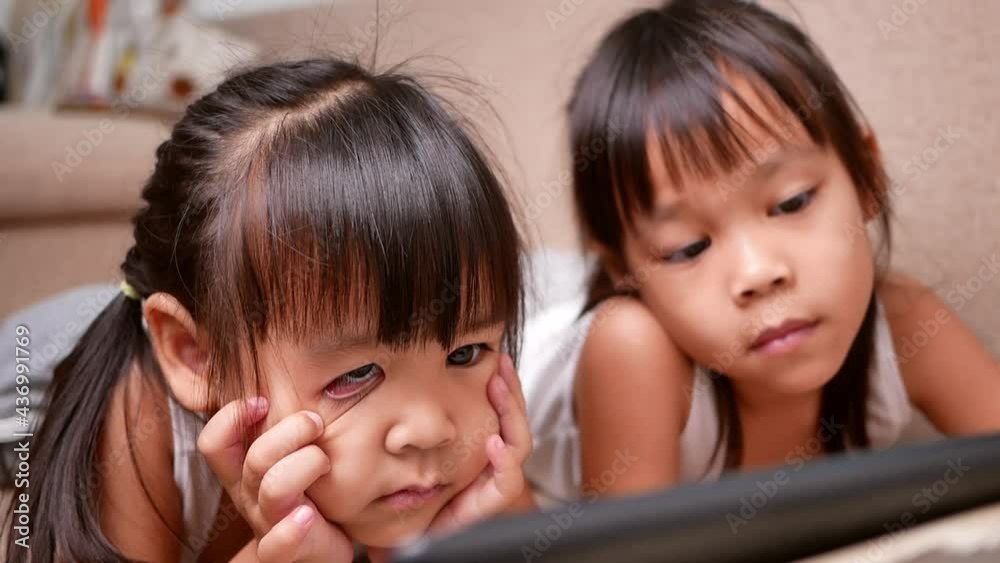 Two sibling girls lying on the sofa and watching digital tablets together in the living room. Two sibling girls lying on the sofa and watching digital tablets together in the living room.