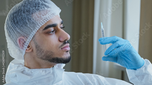 Young arabic male hispanic doctor practitioner surgeon in special medical protective uniform wears rubber gloves holds syringe with medicine vaccine injection for covid-19 anti coronavirus drug remedy