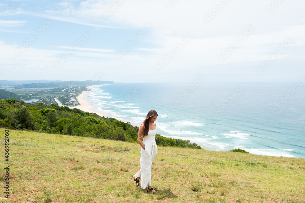 A woman in white walks through a field with the ocean in the background