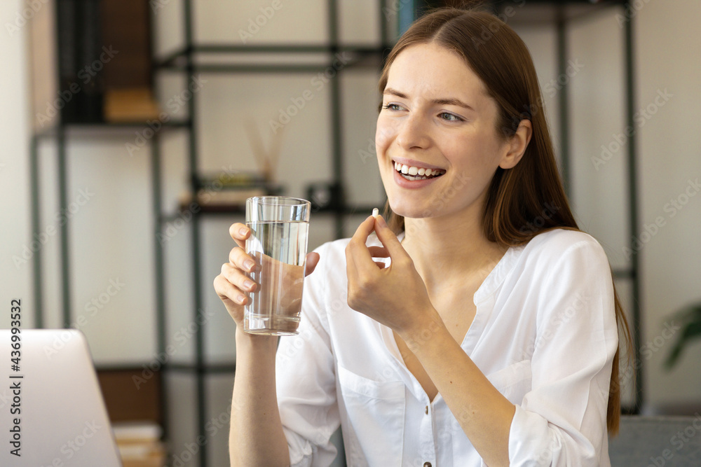 Beautiful young caucasian woman with a glass of water drinks vitamins sitting at home office Beautiful young caucasian woman with a glass of water drinks vitamins sitting at home office