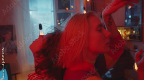 Two young beautiful girlfriends holding incense sticks, dancing back to back and looking at camera while having fun with friends at home retro party