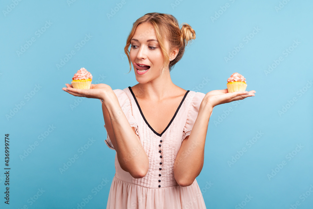 Young adult adorable blonde female with two hair buns wearing dress holding two cakes in hands, showing tongue, wants to lick and eat dessert. Indoor studio shot isolated on blue background.