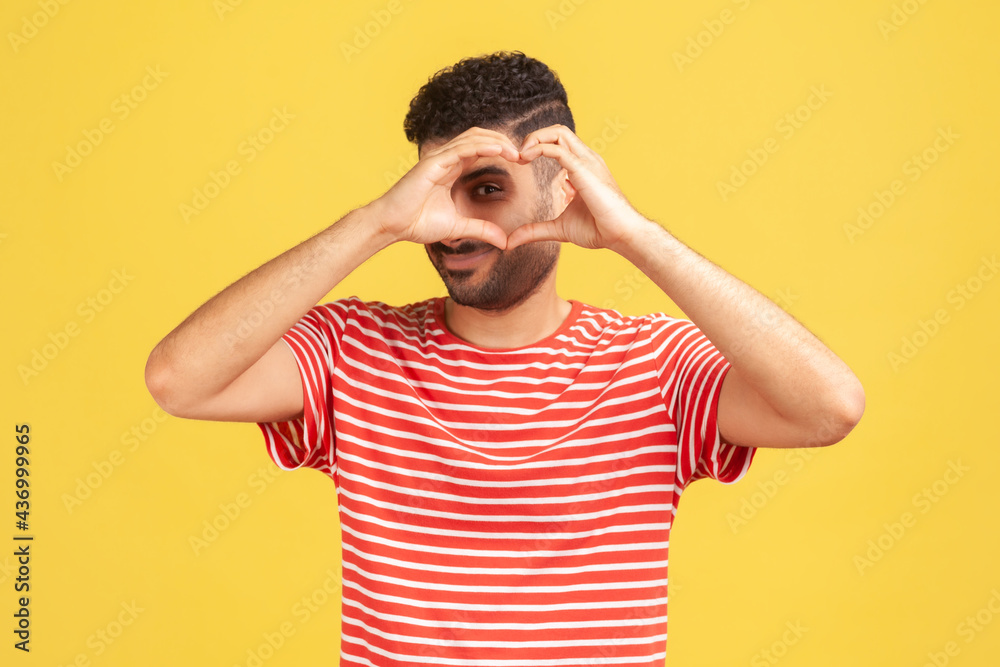 Romantic enamored man with beard in striped t-shirt looking inside heart he made with fingers, showing his feelings, love confession. Indoor studio shot isolated on yellow background