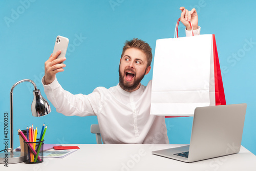 Fotografia Positive bearded man blogger making selfie or recording video, posing at smartphone camera with shopping bags, bragging with purchases