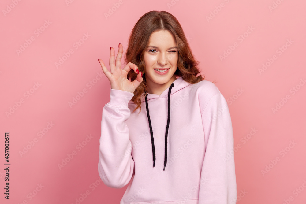 Portrait of satisfied beautiful young woman with brunette hair in hoodie looking at camera showing ok sign gesture and winking. Indoor studio shot, isolated on pink background Portrait of satisfied beautiful young woman with brunette hair in hoodie looking at camera showing ok sign gesture and winking. Indoor studio shot, isolated on pink background
