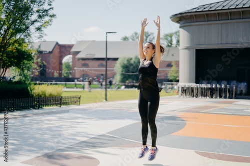Positive active female model has cardio training outdoors jumps high keeps arms raised dressed in activewear being strong fit spends free time on stadium has regular morning workout. Fitness and sport © VK Studio