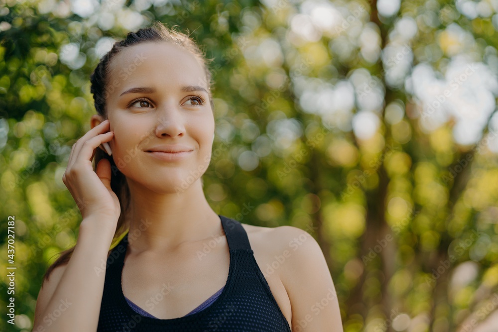 Healthy lifestyle concept. Outdoor shot of sportswoman enjoys favorite playlist in earbuds while training in park has gentle smile concentrated into distance poses over blurred nature background Healthy lifestyle concept. Outdoor shot of sportswoman enjoys favorite playlist in earbuds while training in park has gentle smile concentrated into distance poses over blurred nature background