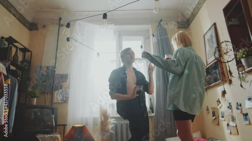 Zoom in shot of young tipsy man and woman drinking and dancing on bed in cozy retro room
