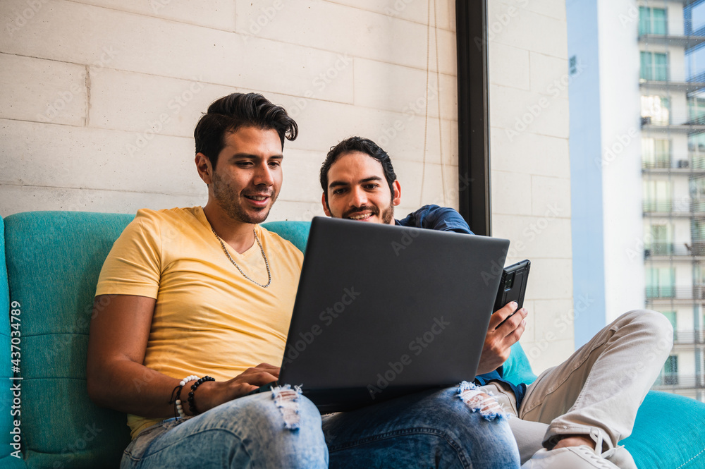 Smiling ethnic gays sharing laptop and smartphone on sofa Smiling ethnic gays sharing laptop and smartphone on sofa
