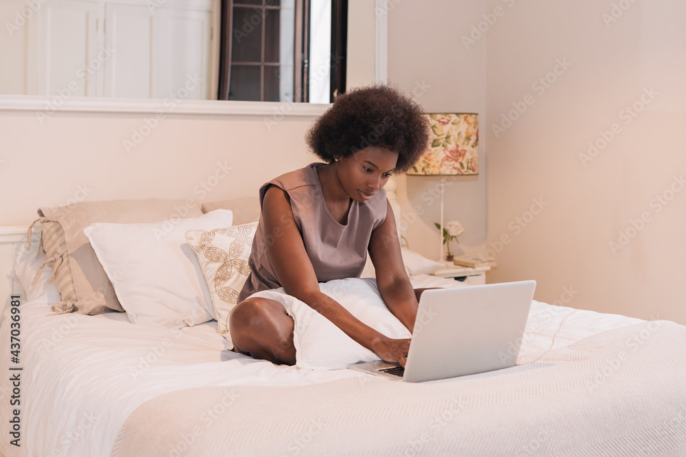 Focused self employed black woman using laptop on bed in modern apartment Focused self employed black woman using laptop on bed in modern apartment