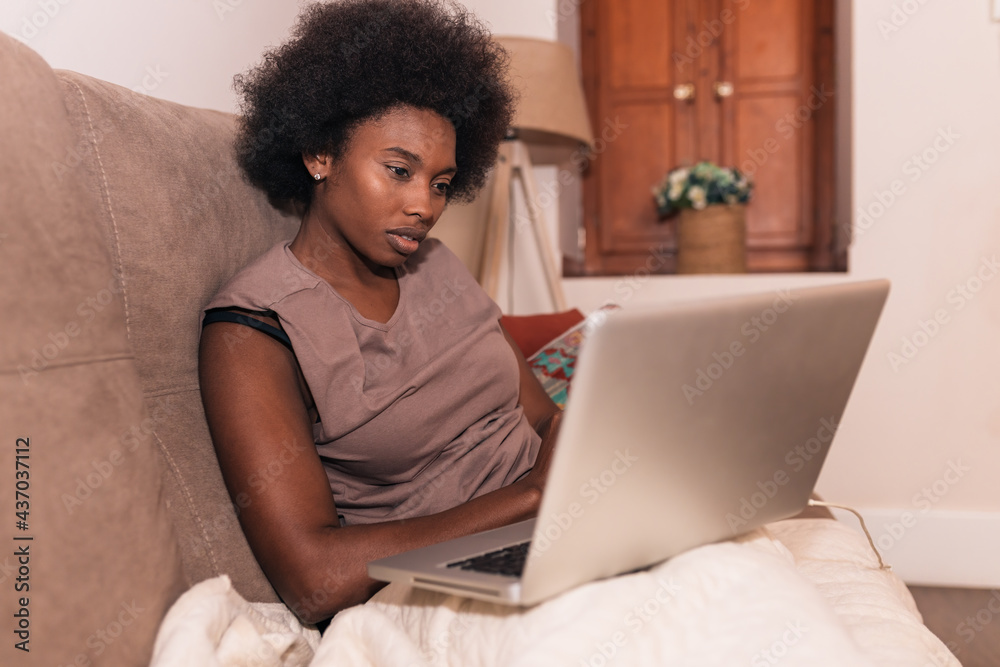 Focused self employed black woman using laptop on bed in modern apartment
