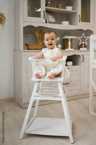 a cheerful little girl in a white dress is sitting in a high white chair in the kitchen. Food for children