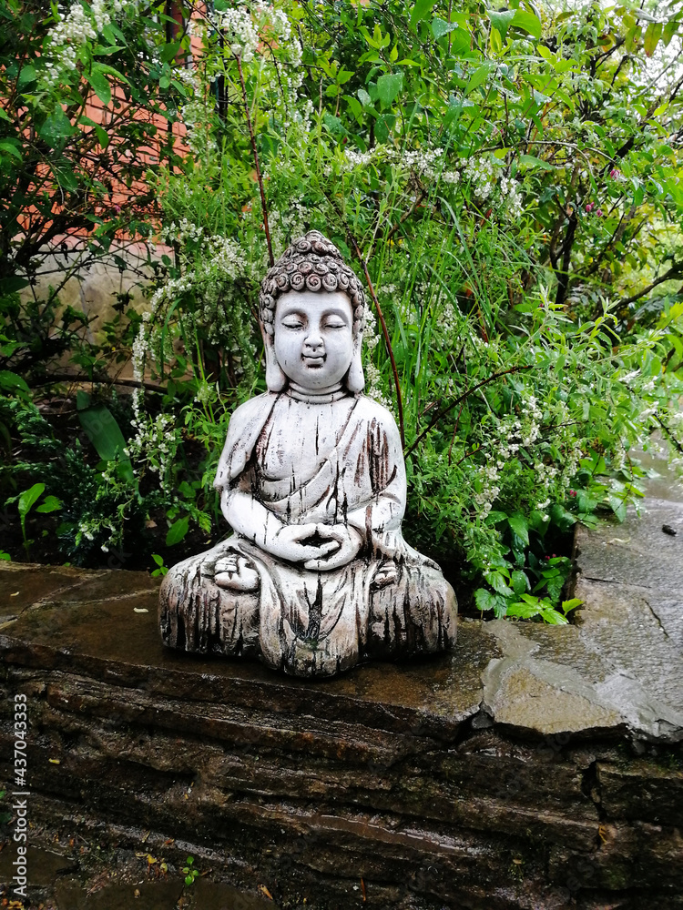 A statue of Buddha, a glazed ceramic Buddha figurine in a spring garden against a background of green leaves.  Mobile phone snapshot.  Illustration for Buddhist and yoga themes.