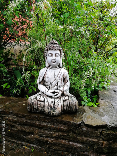A statue of Buddha, a glazed ceramic Buddha figurine in a spring garden against a background of green leaves.  Mobile phone snapshot.  Illustration for Buddhist and yoga themes.
