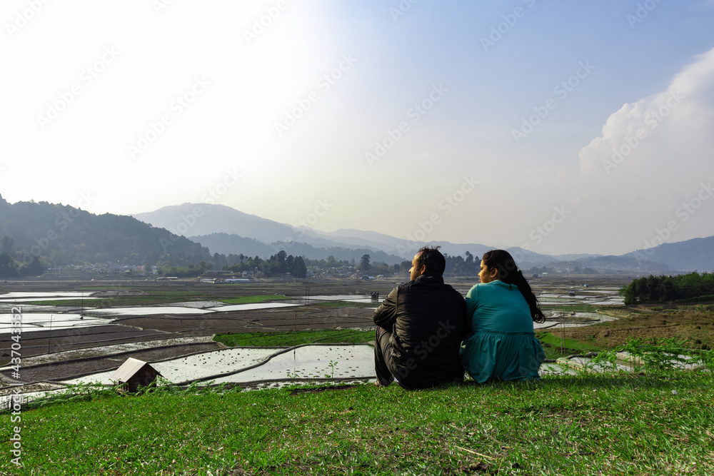 young couple sitting at hill top with paddy fields and mountains in background at evening