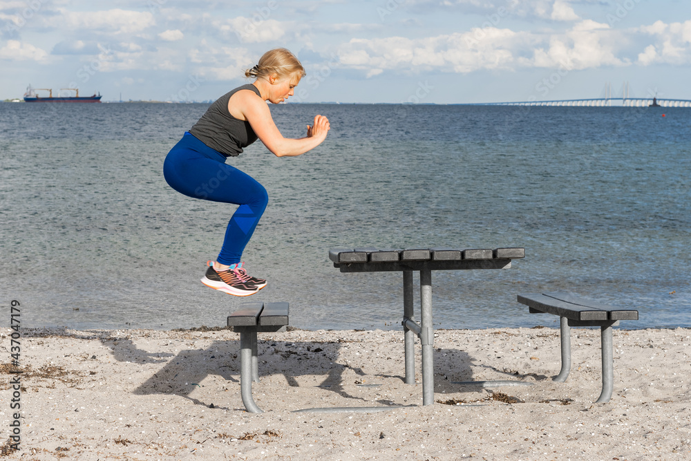 Young caucasian woman doing gym exercises and training on the beach in  Amager, Copenhagen, Denmark. The girl is doing box jumps on a bench. It is  a sunny spring evening. Stock Photo