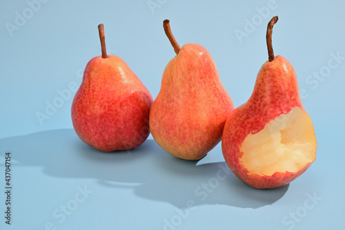 Three yellow-red pears stand side by side with cuttings up. One pear has been bitten off. Fruits cast a harsh shadow