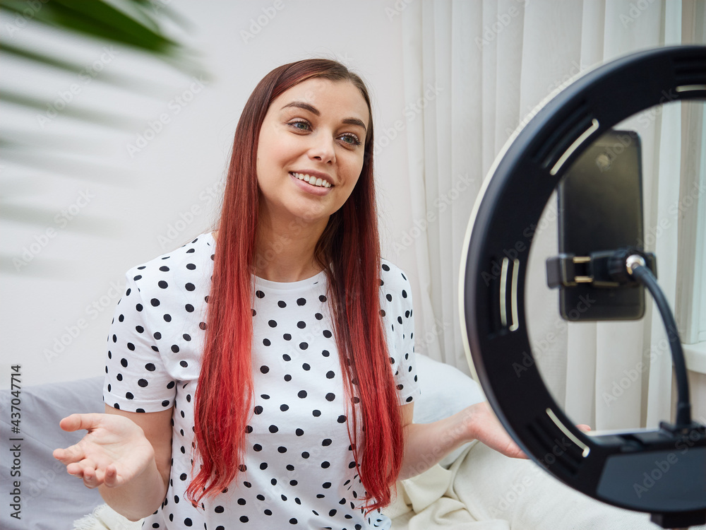A cute female blogger is broadcasting online at home using a ring lamp. The woman communicates with subscribers, gestures with her hands. A cute female blogger is broadcasting online at home using a ring lamp. The woman communicates with subscribers, gestures with her hands.