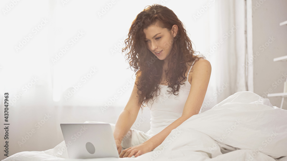 curly young freelancer using laptop while sitting on bed curly young freelancer using laptop while sitting on bed
