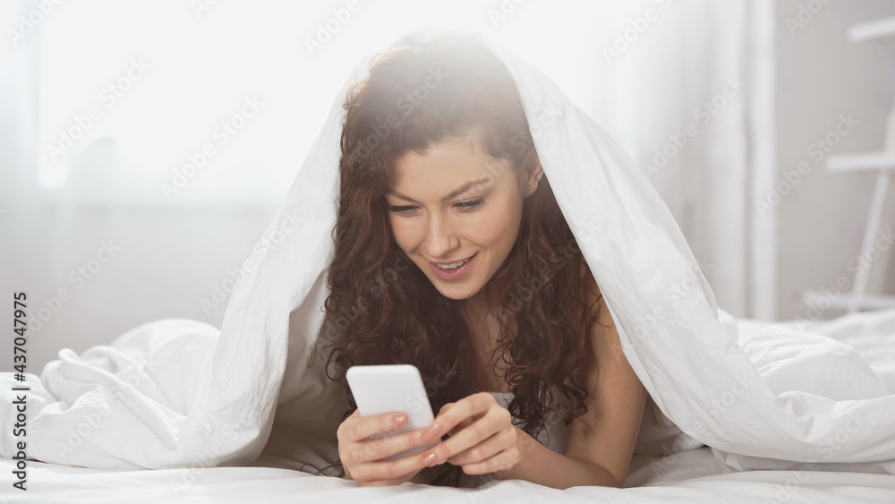 happy young woman using smartphone while lying under blanket