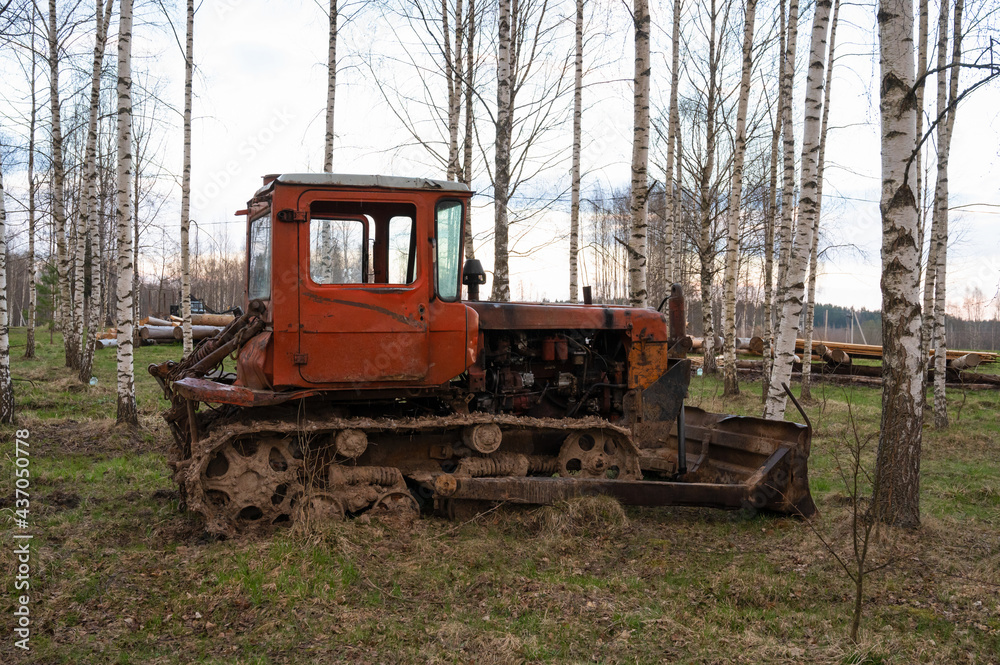 An old rusty red tractor among the trees in the open air on a driverless farm. Agricultural machinery An old rusty red tractor among the trees in the open air on a driverless farm. Agricultural machinery