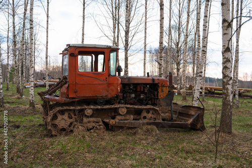 An old rusty red tractor among the trees in the open air on a driverless farm. Agricultural machinery