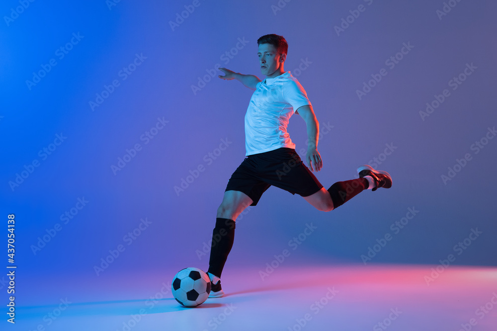 Fototapeta premium Young Caucasian man, male soccer football player training isolated on gradient blue pink background in neon light