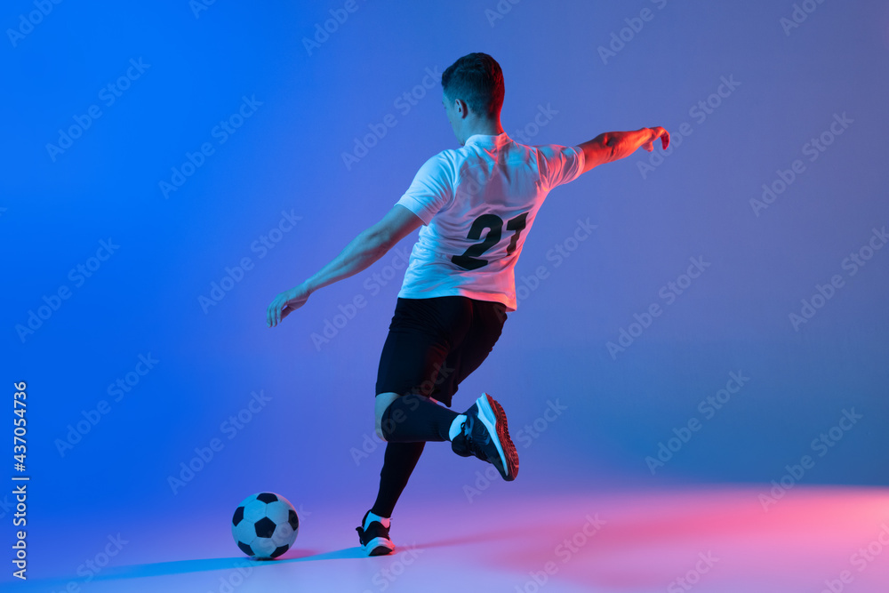 Young Caucasian man, male soccer football player training isolated on gradient blue pink background in neon light. Back view