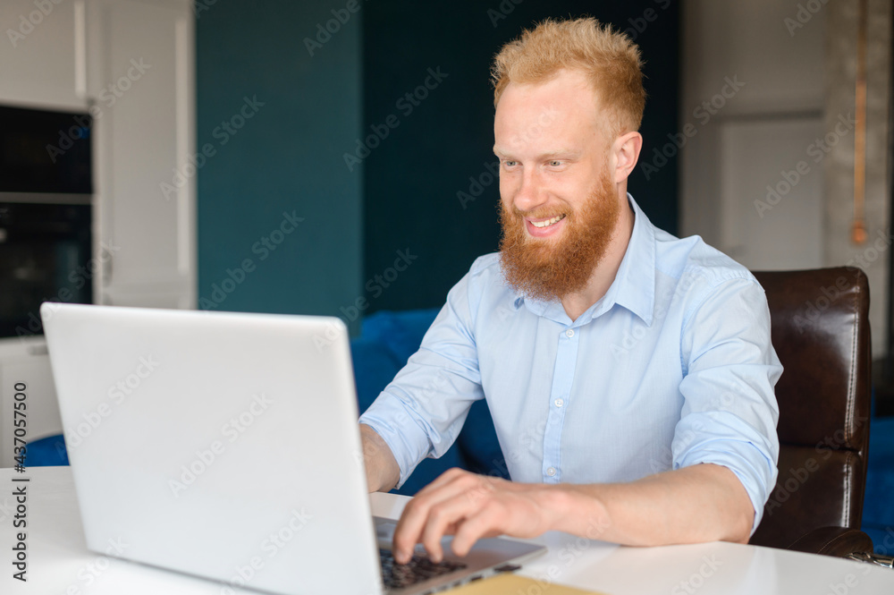 Cheeful red-haired hipster man using laptop computer for remote work, happy young bearded male in formal shirt typing on the keyboard sitting at the desk in loft home office Cheeful red-haired hipster man using laptop computer for remote work, happy young bearded male in formal shirt typing on the keyboard sitting at the desk in loft home office