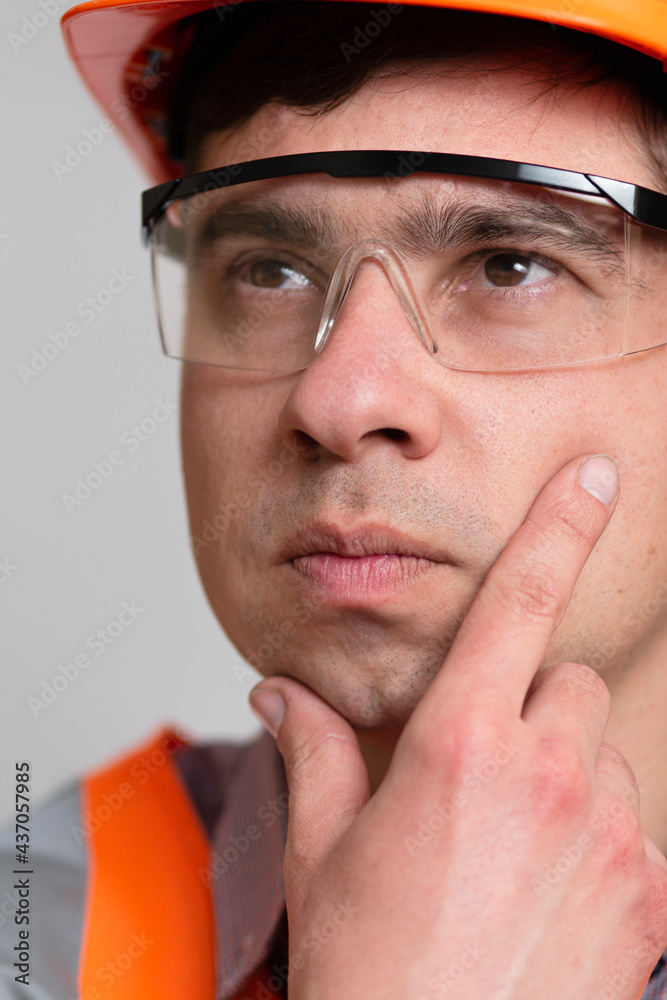 close-up face of thinking young engineer with hand on chin in hard hat and glasses on gray studio background close-up face of thinking young engineer with hand on chin in hard hat and glasses on gray studio background