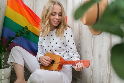 Young millennial hippie woman sitting on balcony play guitar. Music lesson and singer sitting. LGBTQ rainbow flag on background. Online music tuition. Playing instrument