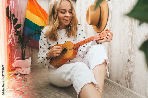 Young millennial hippie woman sitting on balcony play guitar. Music lesson and singer sitting. LGBTQ rainbow flag on background. Online music tuition. Playing instrument