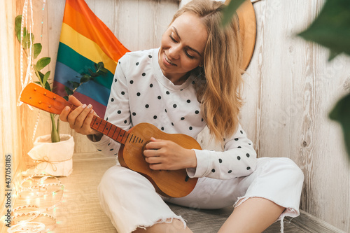Young millennial hippie woman sitting on balcony play guitar. Music lesson and singer sitting. LGBTQ rainbow flag on background. Online music tuition. Playing instrument
